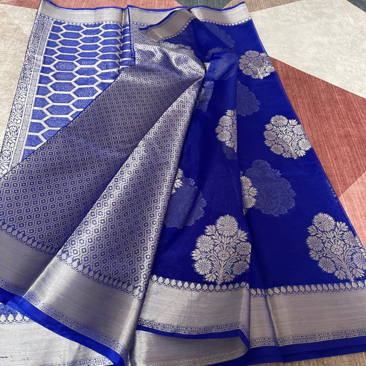 A royal blue silk saree with intricate silver Banarasi zari floral motifs and borders, draped elegantly on a patterned surface.